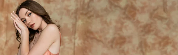 Porträt einer anmutigen jungen Frau mit brünetten Haaren und goldener Halskette, die im rosafarbenen Slip-Kleid auf fleckigem beigem Hintergrund posiert, Sinnlichkeit, Eleganz, Blick in die Kamera, Hände in Gesichtsnähe, Banner — Stockfoto