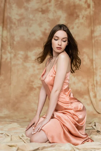 Full length of stunning young woman with brunette hair sitting in sexy slip dress on linen fabric and looking away on mottled beige background, sophistication, sensuality, elegance — Stock Photo