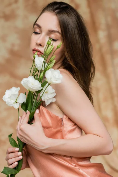 Bezaubernde junge Frau mit brünetten Haaren und geschlossenen Augen, die im Slip-Kleid posiert und weiße Eustoma-Blumen auf fleckigem beigem Hintergrund hält, Sinnlichkeit, Raffinesse, Eleganz — Stockfoto