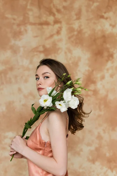 Jeune femme aux cheveux bruns posant en robe glissante rose et soie et tenant des fleurs d'eustomie blanches sur fond beige marbré, sensualité, sophistication, élégance, regardant la caméra — Photo de stock