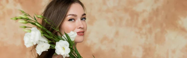Portrait de jeune femme heureuse aux cheveux bruns souriant tout en tenant des fleurs d'eustomie blanches sur fond beige marbré, sensualité, sophistication, élégance, regardant la caméra, bannière — Photo de stock