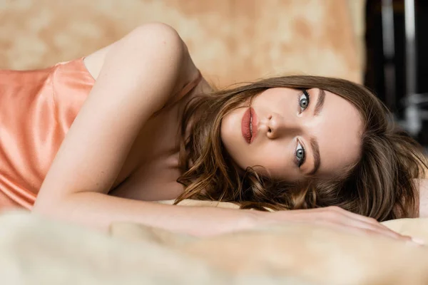 Graceful young woman with brunette hair lying in silk slip dress and looking at camera on blurred and linen fabric on mottled beige background, sophistication,  sensuality, elegance — Stock Photo