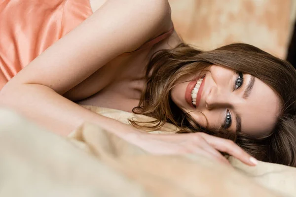 Happy and graceful young woman with brunette hair lying in silk slip dress and looking at camera on blurred and linen fabric on mottled beige background, sophistication, sensuality, elegance — Stock Photo