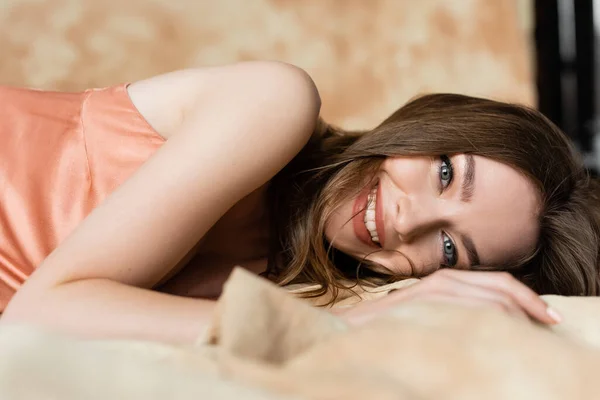 Positive and graceful young woman with brunette hair lying in silk slip dress and looking at camera on blurred and linen fabric on mottled beige background, sophistication, sensuality, elegance — Stock Photo