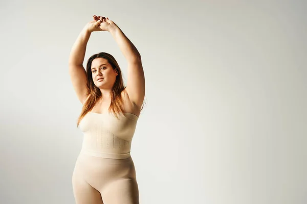 Plus size woman with natural Makeup posing with raised hands in bege strapless top and underwear in studio isolated on grey background, olhando para câmera, corpo positivo, autoconfiança — Fotografia de Stock