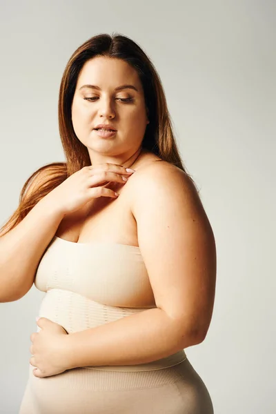 Plus size woman with natural makeup posing in beige strapless top and touching shoulder gently in studio isolated on grey background, looking away, body positive, self-love — Stock Photo