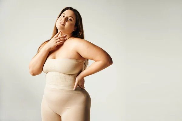 Woman with plus size body touching her neck and looking at camera while posing with hand on hip in beige strapless top and underwear in studio isolated on grey background, body positive, self-love — Stock Photo
