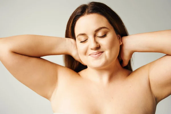 Portrait of pleased woman with plus size body and closed eyes touching hair and posing with bare shoulders isolated on grey background in studio, body positive, self-love — Stock Photo