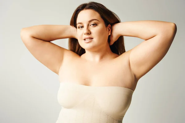 Sensual woman with plus size body touching hair and posing in beige strapless top in studio isolated on grey background, body positive, self-love, looking at camera, self-esteem — Stock Photo