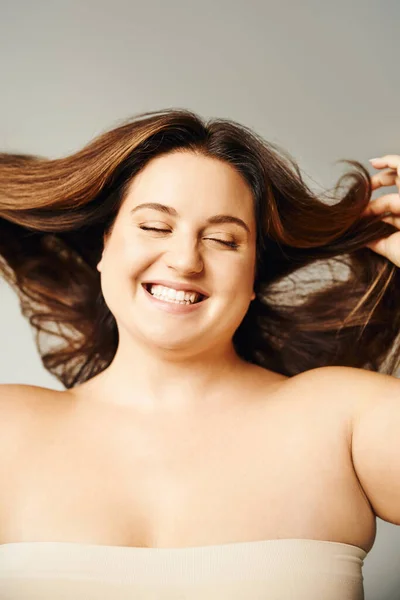 Porträt einer strahlenden Frau mit Plus-Size-Körper und geschlossenen Augen, die Haare berührt und mit nackten Schultern auf grauem Hintergrund im Studio posiert, körperbetont, selbstverliebt — Stockfoto