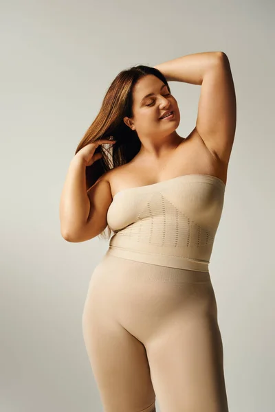Radiant woman with closed eyes touching hair and posing with bare shoulders in underwear isolated on grey background in studio, body positive, self-love, happy plus size — Stock Photo