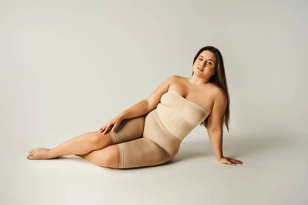Barfüßige und selbstbewusste Frau in trägerlosem Top mit nackten Schultern und Unterwäsche, die im Studio auf grauem Hintergrund posiert, körperbetont, selbstverliebt, plus Größe — Stockfoto