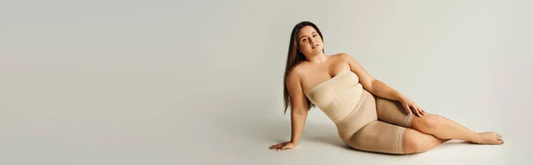 Barfüßige und selbstbewusste Frau in trägerlosem Top mit nackten Schultern und Unterwäsche, die im Studio auf grauem Hintergrund posiert, körperbetont, selbstverliebt, plus Größe, Banner — Stock Photo