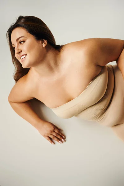 Draufsicht einer lächelnden Frau in trägerlosem Top mit nackten Schultern und Unterwäsche, die im Studio auf grauem Hintergrund posiert, körperbetont, selbstverliebt, plus Größe, Figurentyp, wegschauen — Stockfoto