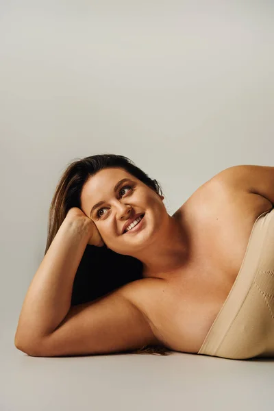 Cheerful woman with plus size body in strapless top with bare shoulders and underwear posing while lying in studio on grey background, body positive, self-love, relaxing, looking away — Stock Photo