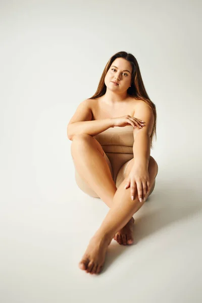 Barfuß-Model mit Plus-Size-Körper in trägerlosem Top mit nackten Schultern und Unterwäsche posiert im Studio auf grauem Hintergrund, körperbetont, selbstverliebt, entspannend, in die Kamera schauend — Stockfoto