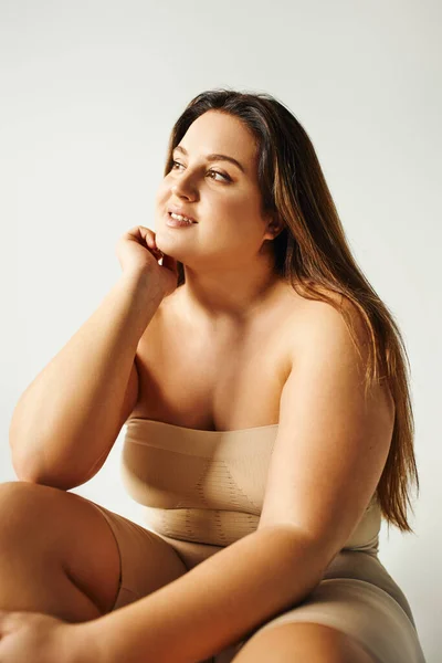 Fröhliches Model mit Plus-Size-Körper in trägerlosem Top mit nackten Schultern posiert, während sie im Studio isoliert auf grauem Hintergrund sitzt, körperbetont, selbstverliebt, entspannend, wegschauend — Stockfoto