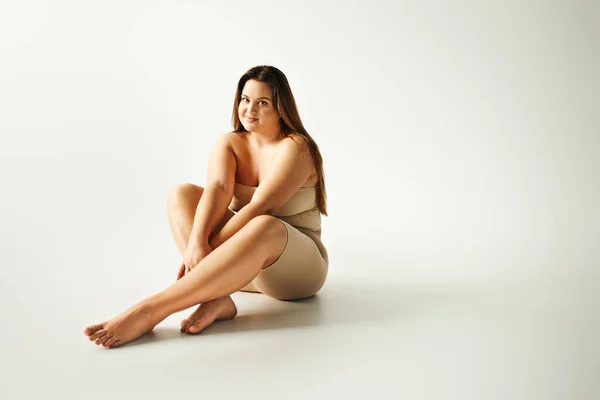 Barefoot and joyous woman with plus size body in strapless top with bare shoulders and underwear sitting in studio on grey background, body positive, self-love, looking at camera, full length — Stock Photo