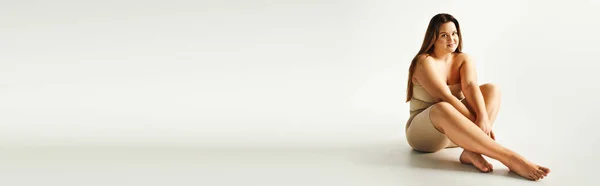 Femme pieds nus et joyeuse avec un corps de taille plus en haut sans bretelles avec des épaules nues et des sous-vêtements posant en studio sur fond gris, corps positif, regardant la caméra, bannière — Photo de stock