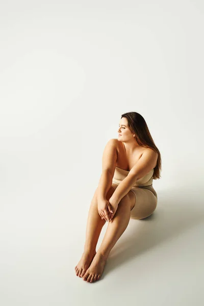 Full length of barefoot and confident woman with plus size body in strapless top with bare shoulders and underwear posing while sitting in studio on grey background, body positive, figure type — Stock Photo