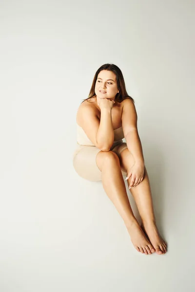 Volle Länge der verträumten Plus-Size-Frau in trägerlosem Top mit nackten Schultern und Unterwäsche posiert, während sie im Studio auf grauem Hintergrund sitzt, körperbetont, Figurentyp, wegschauend — Stockfoto