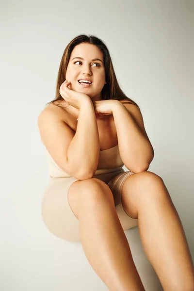 Dreamy plus size woman in strapless top with bare shoulders and underwear posing while sitting in studio on grey background, body positive, figure type, smiling while looking away — Stock Photo
