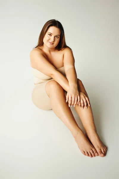 Piena lunghezza di piedi nudi e bella donna con corpo plus size in top senza spalline con spalle nude e biancheria intima in posa mentre seduto in studio su sfondo grigio, corpo positivo, tipo di figura — Foto stock