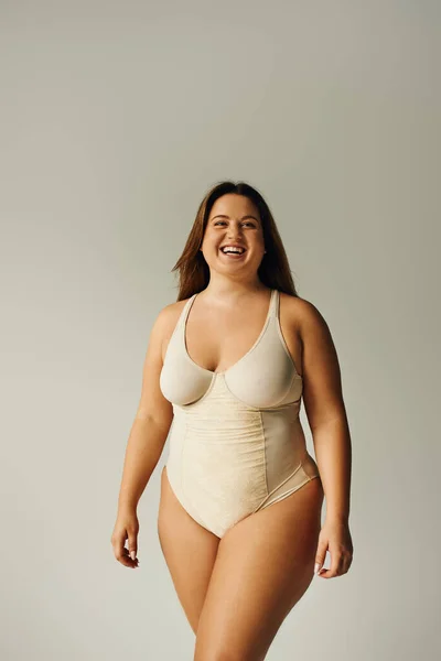 Happy plus size woman in beige bodysuit posing while standing in studio on grey background, body positive, figure type, self-esteem, smiling while looking at camera — Stock Photo