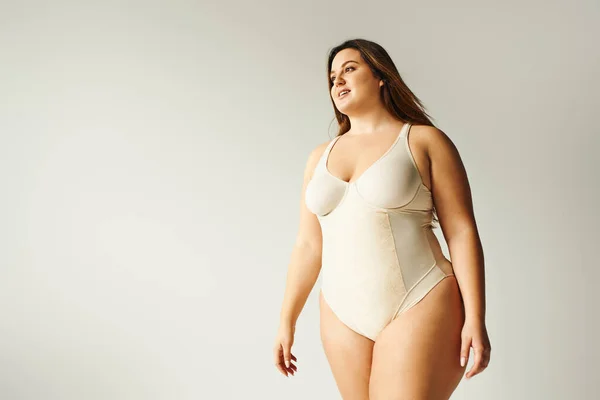 Happy plus size woman in beige bodysuit posing while standing in studio on grey background, body positive, figure type, self-esteem, smiling while looking away — Stock Photo