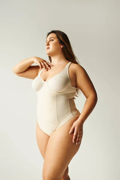 Curvy woman with natural makeup posing in beige bodysuit while standing in studio on grey background, body positive, figure type, self-esteem, smiling while looking away — Stock Photo
