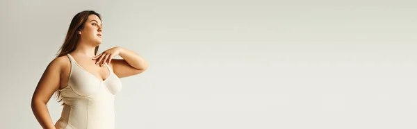 Kurvige Frau mit natürlichem Make-up posiert im beigen Body-Anzug im Studio auf grauem Hintergrund, körperbetont, Figurentyp, Selbstwertgefühl, lächelt beim Wegschauen, Banner — Stockfoto