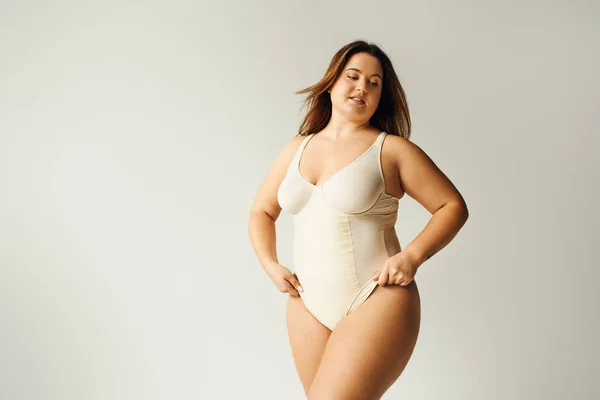 Tattooed and brunette curvy woman with plus size body posing in beige bodysuit while standing in studio on grey background, body positive, figure type, self-esteem, tattoo translation: harmony — Stock Photo