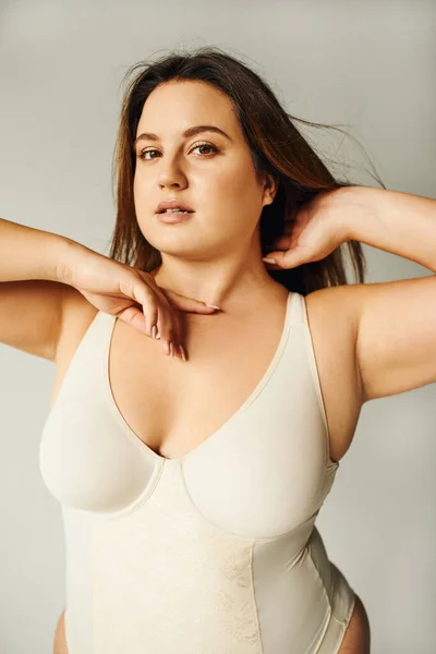 Portrait of brunette curvy woman with plus size body posing in beige bodysuit while standing and touching hair in studio on grey background, body positive, figure type, looking at camera — Stock Photo