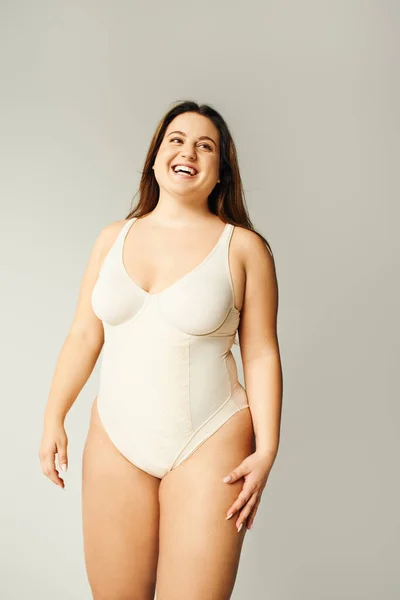 Portrait of positive and curvy woman with plus size body posing in beige bodysuit while laughing on grey background, body positive, figure type, looking away while standing in studio — Stock Photo