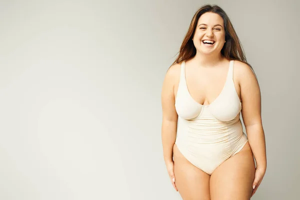 Portrait of happy and curvy woman with plus size body posing in beige bodysuit while laughing on grey background, body positive, figure type, looking at camera while standing in studio — Stock Photo