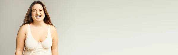 Retrato de mulher feliz e curvilínea com corpo plus size posando em bodysuit bege enquanto rindo em fundo cinza, corpo positivo, tipo de figura, olhando para a câmera enquanto estava em estúdio, banner — Fotografia de Stock