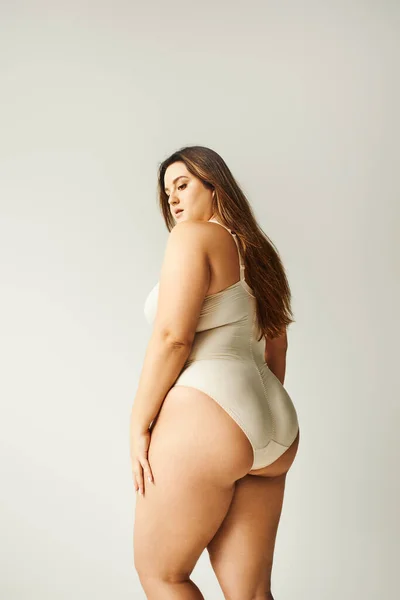Brunette and curvy woman wearing beige bodysuit and standing isolated on grey background, self-confidence, figure type, looking down, body positivity movement — Stock Photo