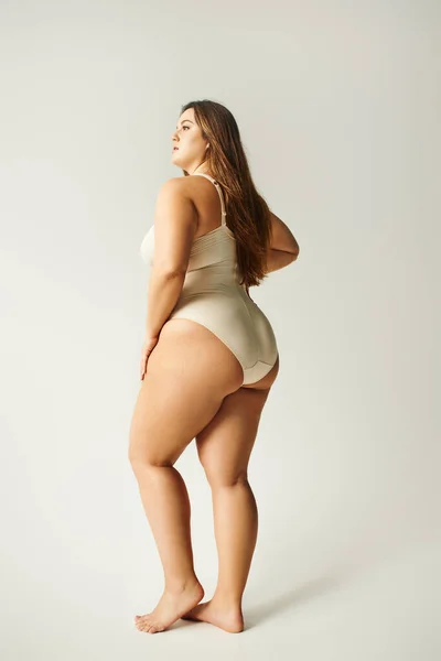 Full length of brunette and curvy woman wearing beige bodysuit and standing with bare feet on grey background, body positive, figure type, looking away, body positivity movement — Stock Photo