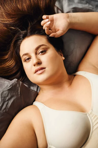 Top view of attractive and curvy woman wearing beige bodysuit and lying on bed with grey bedding while looking at camera, body positive, figure type, modern apartment, portrait — Stock Photo