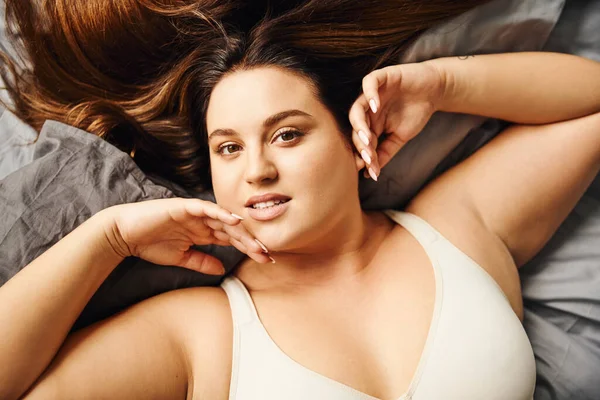 Top view of tattooed plus size woman with natural makeup lying in beige bodysuit on bed with grey bedding while touching face and looking at camera, body positive, modern apartment, portrait — Stock Photo