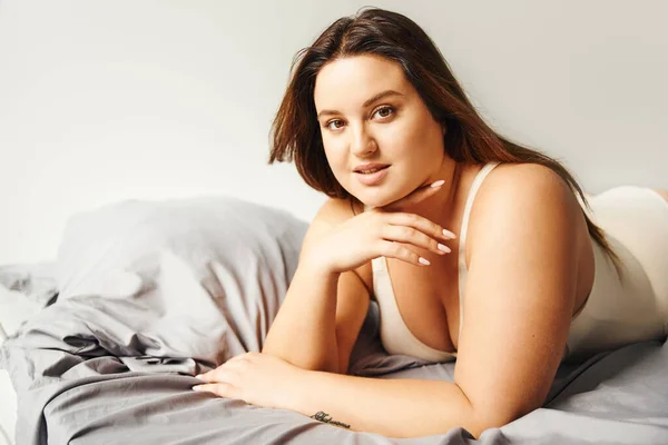 Brunette plus size woman with natural makeup wearing beige bodysuit and resting on bed with grey bedding while looking at camera, body positive, figure type, tattoo translation: harmony — Stock Photo