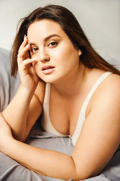 Portrait of attractive plus size woman with natural makeup wearing beige bodysuit and resting on bed with grey bedding while looking at camera, body positive, figure type, modern apartment — Stock Photo