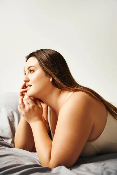 Dreamy plus size woman with natural makeup wearing beige bodysuit and resting on bed with grey bedding while looking away, body positive, figure type, self-confidence — Stock Photo