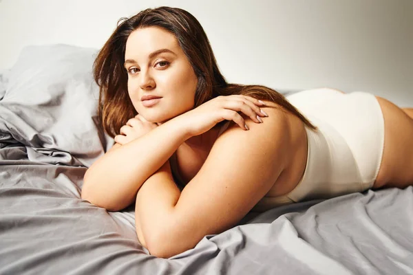 Brunette woman with plus size body and natural makeup wearing beige bodysuit and lying on bed with crossed arms while looking at camera, figure type, body diversity — Stock Photo