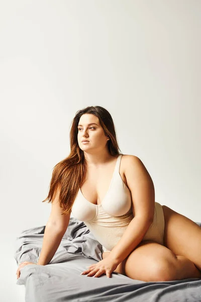 Brunette plus size woman with natural makeup and long hair wearing beige bodysuit and posing on bed with grey bedding while looking away, body positive, figure type, caucasian — Stock Photo