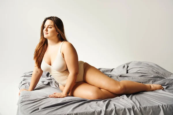 Dreamy brunette plus size woman with natural makeup wearing beige bodysuit and posing on bed with grey bedding while looking away, body positive, figure type, tattoo translation: harmony — Stock Photo