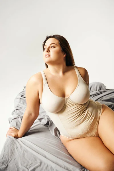 Dreamy and brunette woman with natural makeup and plus size body wearing beige bodysuit and posing on bed with grey bedding, body positive, figure type, looking away — Stock Photo