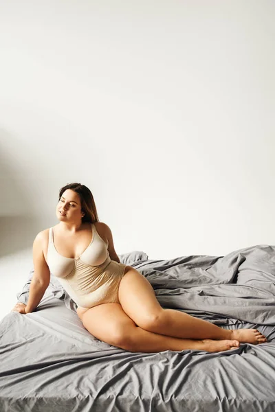 Dreamy and brunette woman with natural makeup and plus size body wearing beige bodysuit and posing on bed with grey bedding, body positive, figure type, bare feet, looking away — Stock Photo