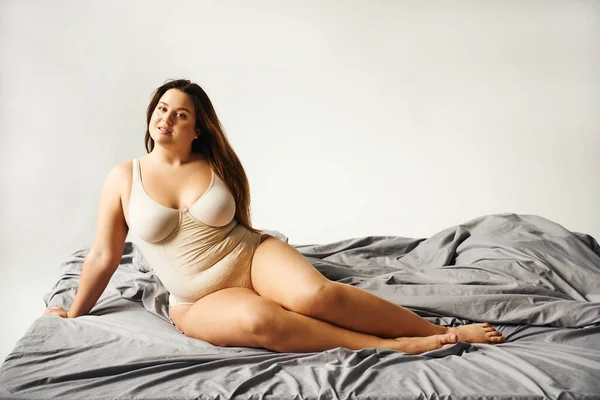 Charmante Frau mit natürlichem Make-up und Plus-Size-Körper trägt beigen Body und posiert auf dem Bett mit grauer Bettwäsche, Körper positiv, Figurentyp, nackte Füße, Blick in die Kamera — Stockfoto