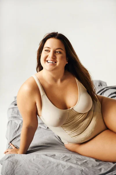 Lächelnde und brünette Frau mit natürlichem Make-up und Plus-Size-Körper im beigen Body und posiert auf dem Bett mit grauer Bettwäsche, körperbetont, Figurentyp, wegschauend — Stockfoto